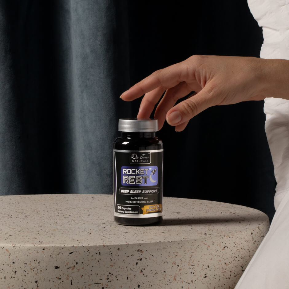 A hand reaching for a bottle of Dr. Jones&rsquo; Naturals Rocket Rest sleep support on a bedside table