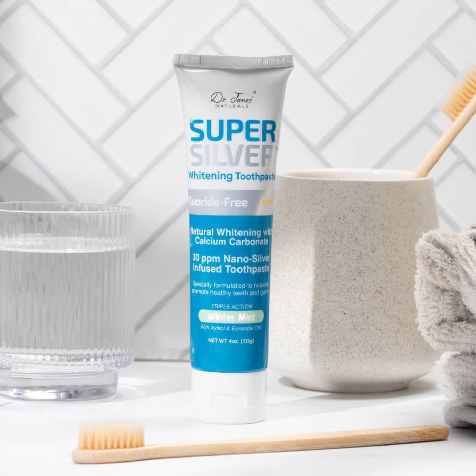 A tube of Dr. Jones&rsquo; Naturals Super Silver whitening toothpaste on a bathroom counter with toothbrushes and a glass of water